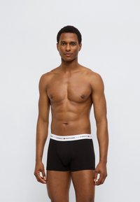 Μαύρα βαμβακερά μποξεράκια με λευκή ελαστική μέση που φέρει το λογότυπο "TOMMY HILFIGER", εμφανιζόμενα σε ένα άτομο με μυώδη σωματότυπο.