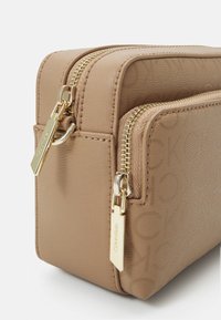 Borsa in pelle sintetica color taupe con dettagli dorati, dotata di due cerniere. Un motivo a logo in rilievo ricopre la superficie, aggiungendo texture e dettaglio.