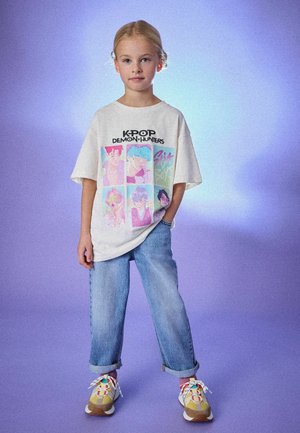 Weißes, übergroßes T-Shirt mit K-Pop-Grafikdruck, blaue Jeans aus Denim und bunte Sneakers mit gemusterten Schnürsenkeln. Der Hintergrund ist in sanftem Lila gehalten.