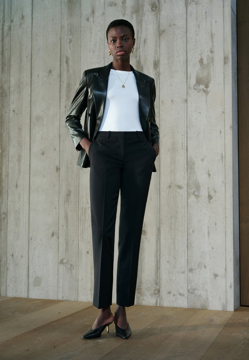Schwarze Leder-Blazer, weißes enges Top, maßgeschneiderte schwarze Hosen und schwarze Absatzschuhe. Glatte Texturen vor einem Betond Hintergrund.