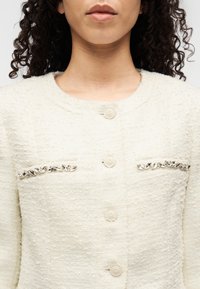 Λευκό υφασμάτινο σακάκι με στρογγυλή λαιμόκοψη, που διαθέτει διακοσμητικές κρυστάλλινες λεπτομέρειες στις τσέπες και στρογγυλά κουμπιά στο εμπρόσθιο μέρος.