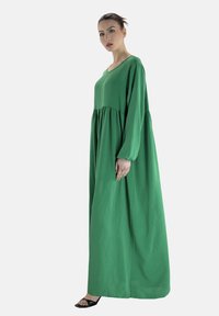 Długa zielona sukienka z miękkiego materiału, o luźnym kroju, z długimi rękawami i marszczoną talią. W połączeniu z czarnymi sandałami.