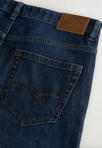 Ciemnoniebieskie dżinsy wykonane z denimu, posiadające prostokątną łatkę z logo na pasie oraz przeszywaną tylną kieszeń z ozdobnym wzorem.