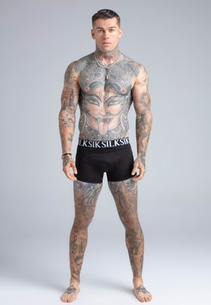 Muskularny mężczyzna z rozległymi tatuażami, w tym dużą twarzą na torsie, ubrany w czarne szorty, stojący boso na jednolitym szarym tle.