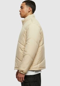 Beige dunjakke med høy krage, cropped design, kviltet tekstur, elastiske mansjetter og glidelås. Brukt over en hvit skjorte.