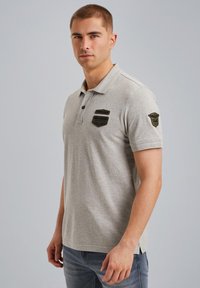 Camisa polo gris con dos parches negros en el pecho y el hombro; mangas cortas, cuello abotonado y aberturas laterales.