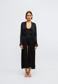 Čierny saténový župan s čipkovými detailmi, dlhými rukávmi a opaskom na zaväzovanie, nosený cez čierne body. Hladká textúra, plynulý strih, dĺžka po kolená.