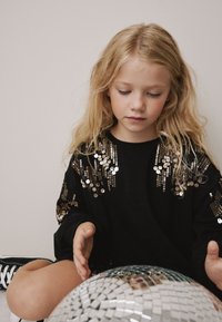 Črna majica z dolgimi rokavi, okrašena z zlato bleščicami. Otrok sedi z ogledalom disco kroglo in gleda navzdol.