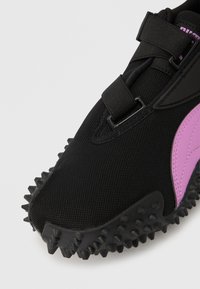 Μαύρο αθλητικό παπούτσι με υφασμάτινο επάνω μέρος με υφή, μοβ λεπτομέρειες και εξωτερική σόλα από καουτσούκ με καρφιά για πρόσφυση. Ρυθμιζόμενο κλείσιμο με λουράκι.