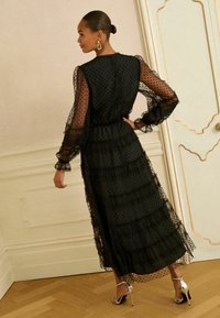 Rochie neagră cu buline albe, cu volane suprapuse, mâneci lungi din material semi-transparent, talie elastică și decupaj în spate. Asortată cu pantofi cu toc înalt argintii.