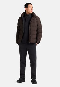 Giacca a vento marrone scuro, con colletto alto e zip, indossata sopra una felpa nera. Abbinata a pantaloni neri e sneakers.