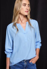 Blouse bleu clair avec un décolleté en V, manches longues avec poignets élastiques, et texture douce et lisse, assortie à un jean en denim.