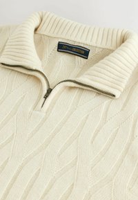 Cream-colored pullover with a ribbed collar, featuring a front zipper and cable knit texture throughout. Brand label positioned at the collar.