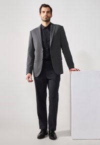 Grå kavaj med slagkrage, två knappar, kombinerad med en svart skjorta och mörka byxor. Svarta läderskor fulländar outfiten.