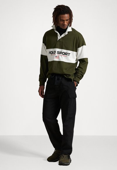 Olijfgroene polo met lange mouwen met een witte streep en een rode "POLO SPORT" logo, gecombineerd met zwarte cargo pants en donkere schoenen.