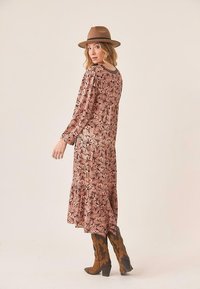 Vestido largo de estampado floral en rosa claro con mangas largas y un dobladillo con volantes. Combinado con botas marrones de caña alta con estampado y un sombrero de ala ancha.