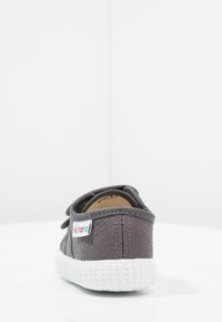 Sneaker in tela grigia con suola in gomma bianca, vista posteriore. Presenta un design cucito e un piccolo logo laterale.