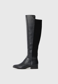 Bota de cuero negra hasta la rodilla con panel trasero de tela elástica, tacón bajo en bloque y pequeño logo metálico de la marca cerca del borde superior.