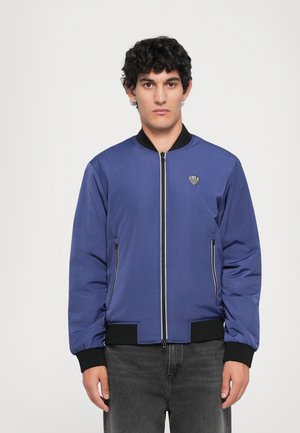 Young man with short curly hair wears a blue zip-up bomber jacket with black cuffs and collar, standing against a plain background.