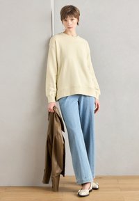 Felpa oversized color beige chiaro con orlo a costine, abbinata a jeans a gamba larga blu chiaro. Il modello tiene in mano una giacca di pelle marrone e indossa scarpe senza lacci.