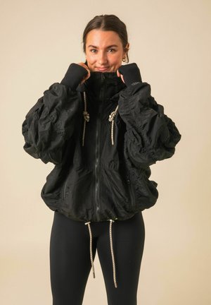 Jeune femme portant une veste noire oversize avec cordons et des leggings noirs, remontant le col tout en souriant à la caméra.