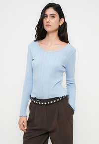 Ανοιχτό μπλε ριπ μακρυμάνικο τοπ με φαρδύ ντεκολτέ και κυματιστές άκρες, σε συνδυασμό με καφέ ψηλόμεσα παντελόνια και ζώνη με κυκλικά καρφιά.