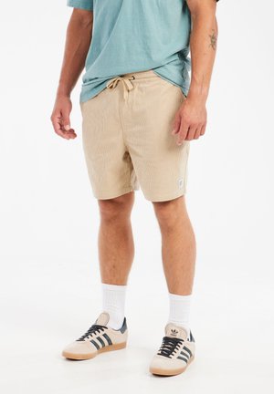 Beige corduroy shorts met een elastische tailleband en een trekkoord. Gepaard met een lichtblauw T-shirt en beige sneakers met zwarte accenten.