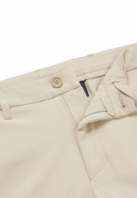 Beiga byxor med knappstängning och dragkedja, med en slät tygtextur och bälteshällor i midjan. Inuti fodrade med kontrasterande material.