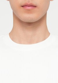 Κάτω μέρος του προσώπου και ο λαιμός ενός ατόμου που φοράει λευκό ριμπ πλεκτό μπλουζάκι με στρογγυλή λαιμόκοψη, με φόντο ένα απλό λευκό τοίχο.