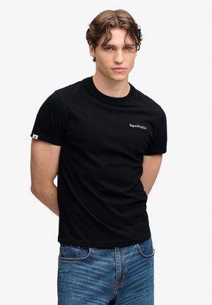Joven con pelo corto y ondulado lleva una camiseta negra lisa de Superdry y vaqueros azules, de pie con las manos detrás de la espalda sobre un fondo claro.