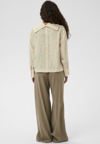 Svetlo rumena bluzica s cvetnim vzorcem, oversize ovratnikom in gumbom na hrbtni strani. V kombinaciji z ohlapenimi bež hlačami.