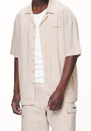 Camisa de botones de manga corta color beige claro con un ajuste relajado, que presenta un pequeño detalle bordado y una textura suave y lisa.
