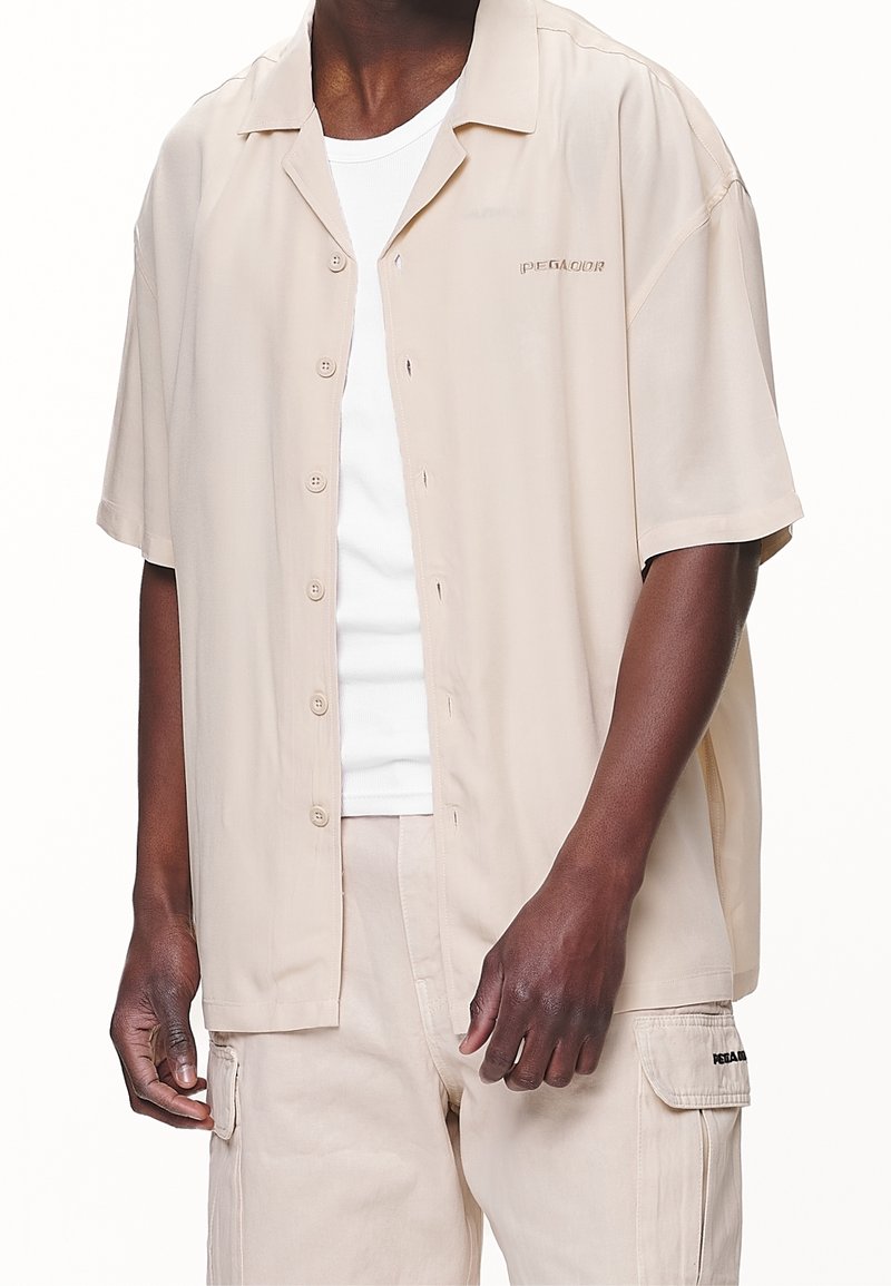 Ljusbeige kortärmad skjorta med knapplist och avslappnad passform, med en liten broderad detalj och mjuk, slät textur.