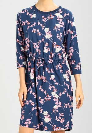 Navyblå kjole med pink og hvide blomsterprint, treskåret ærmer og indsnævret talje, båret af en stående person.