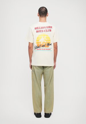 Camiseta corta de manga en crema con un gráfico colorido en la parte de atrás que muestra palmeras y texto. Se lleva con pantalones de rayas claros y zapatos negros.