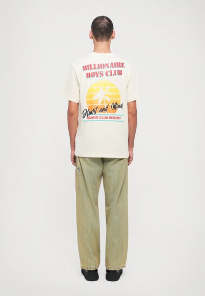 Cream short-sleeve T-shirt featuring a colorful graphic print on the back with palm trees and text. Worn with light striped pants and black shoes.