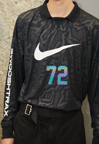 Personne portant une chemise à manches longues noire à motifs avec un grand logo Nike blanc, un numéro 72 holographique, et le texte "Swooshtrax" sur la manche.
