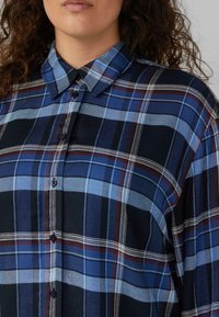 Woman wearing a blue, black, and burgundy plaid button-up shirt with a collar, shown from shoulders to chin with curly hair visible.