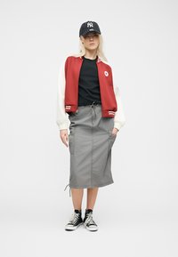 Γυναίκα που φοράει ένα κόκκινο και λευκό varsity μπουφάν, μαύρο t-shirt, γκρι μίντι φούστα, μαύρα παπούτσια ψηλά sneakers και μαύρο καπέλο baseball με ρίγες.