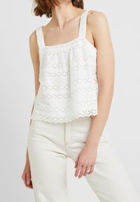 Top blanco sin mangas con patrón de bordado en forma de ojo y dobladillo con volantes, y tirantes anchos. Tejido ligero con un ajuste relajado.