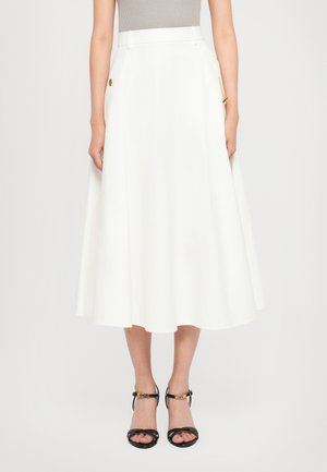 Mujer vestida con una falda midi blanca que tiene un detalle de cadena lateral y sandalias de tacón alto negras con tiras y acentos dorados en forma de letras.