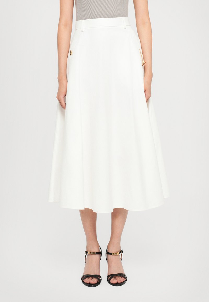 Woman wearing a white midi skirt with a side chain detail and black strappy high-heeled sandals with gold letter accents.