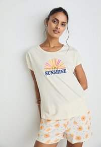 Cream-colored cotton t-shirt featuring a sun graphic with "Hey SUNSHINE" text. Paired with white shorts adorned with yellow and pink sun patterns.