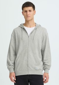 Sweat à capuche gris à fermeture éclair avec cordons, doté d'une poche avant et de poignets côtelés. Porté sur un t-shirt blanc, sur un fond clair.