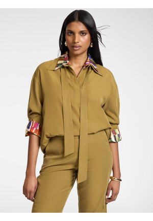 Kobieta ubrana w bluzkę i spodnie w kolorze musztardowym, bluzka ma kolorowy kołnierzyk i mankiety, długie czarne włosy, stoi na jednolitym tle.