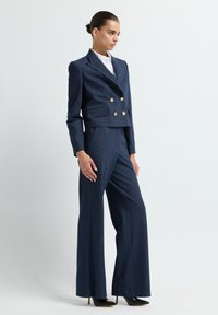 Vrouw in een marineblauwe pinstripe double-breasted blazer met gouden knopen en bijpassende wide-leg broek, staande in profiel tegen een witte achtergrond.