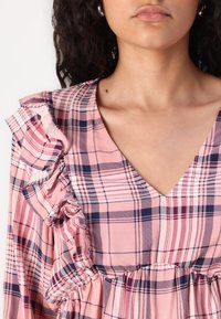 Pink plaid dress featuring a V-neck and ruffled shoulders. Made of soft fabric with white and navy checks. Gathered waist detail.