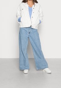 Vit denimjacka med knappstängning, ljusblå tröja under, vida ljusblå jeans och vita sneakers. Enfärgad bakgrund.