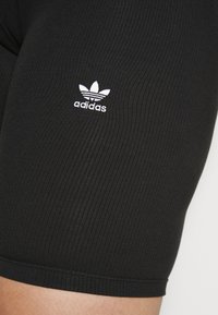 Černé žebrované kraťasy z látky s elastickým pasem. Mají bílý vyšívaný logo Adidas poblíž lemu.