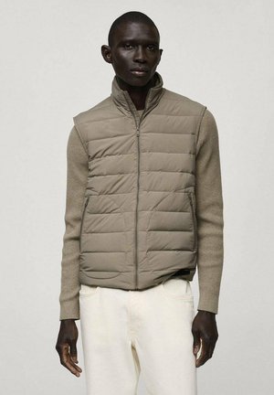 Khaki puffer vest with quilted pattern, zip closure, and faux-zipper pockets, layered over a light-toned, textured long-sleeve top.